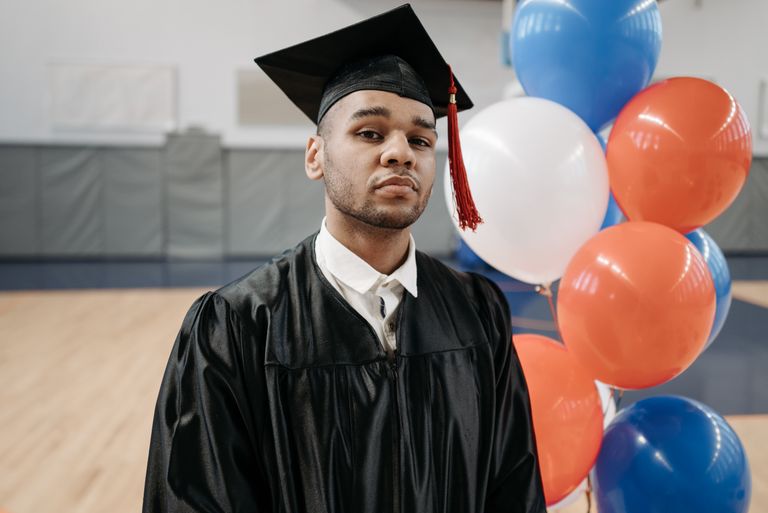 Proud graduate in cap and gown with colorful balloons in a gym setting.