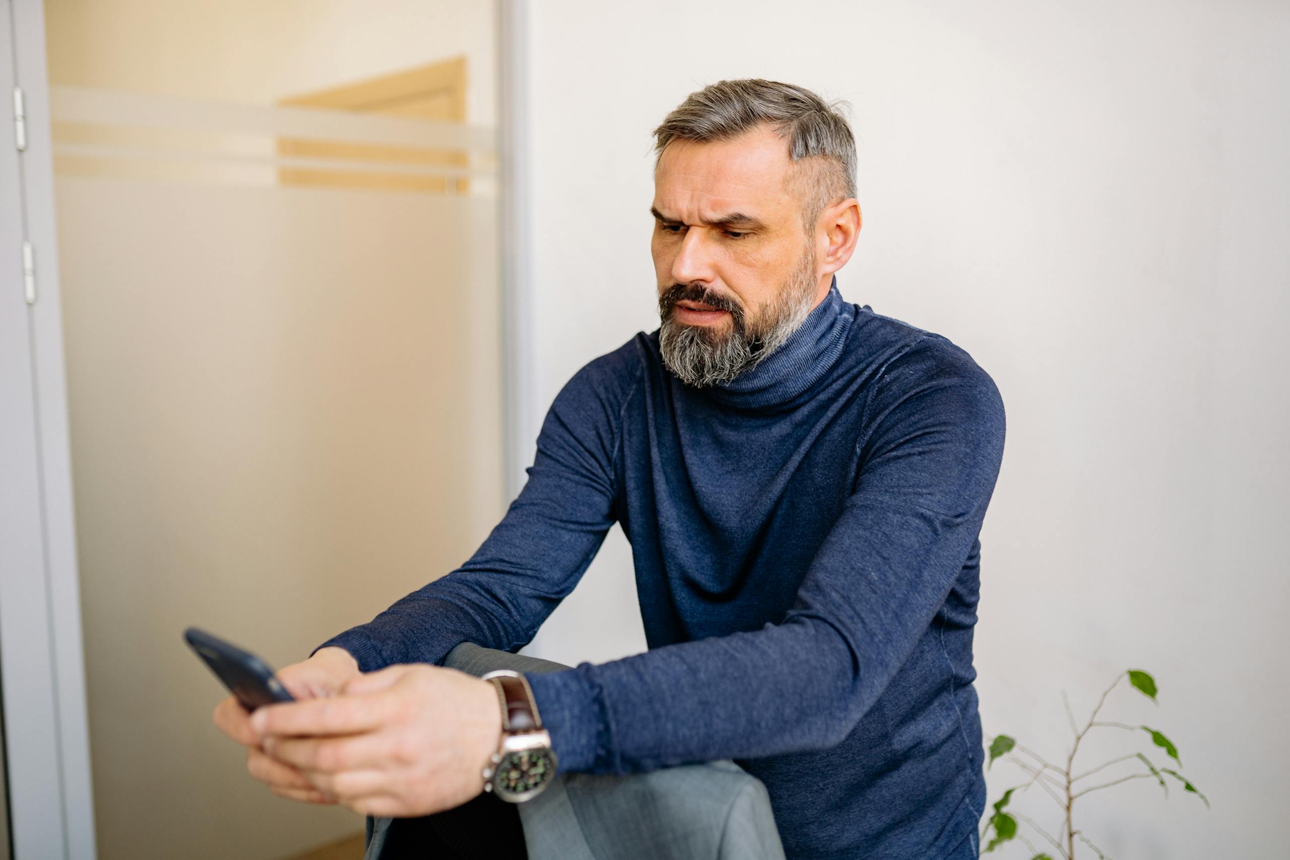 Serious middle-aged man sitting and using a smartphone indoors.
