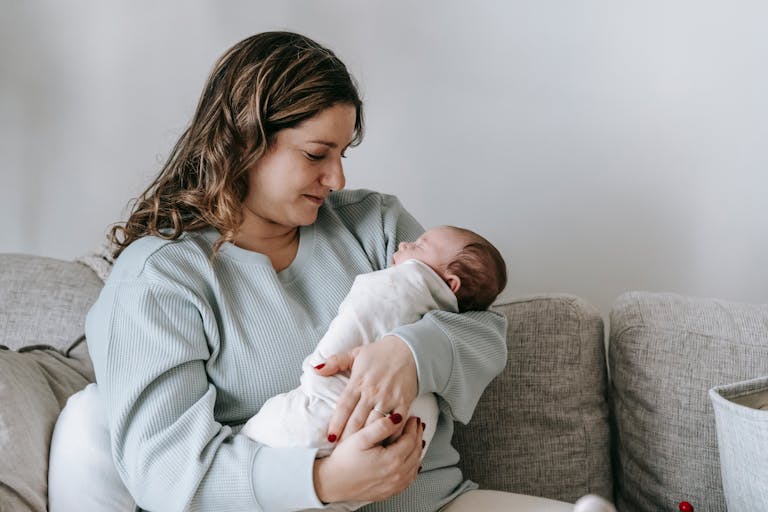 Side view of caring mother with infant baby in arms sitting on comfortable couch in light living room at home