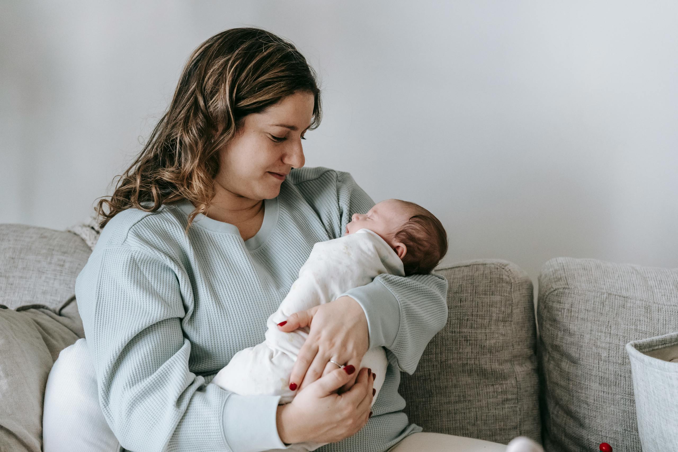 Side view of caring mother with infant baby in arms sitting on comfortable couch in light living room at home