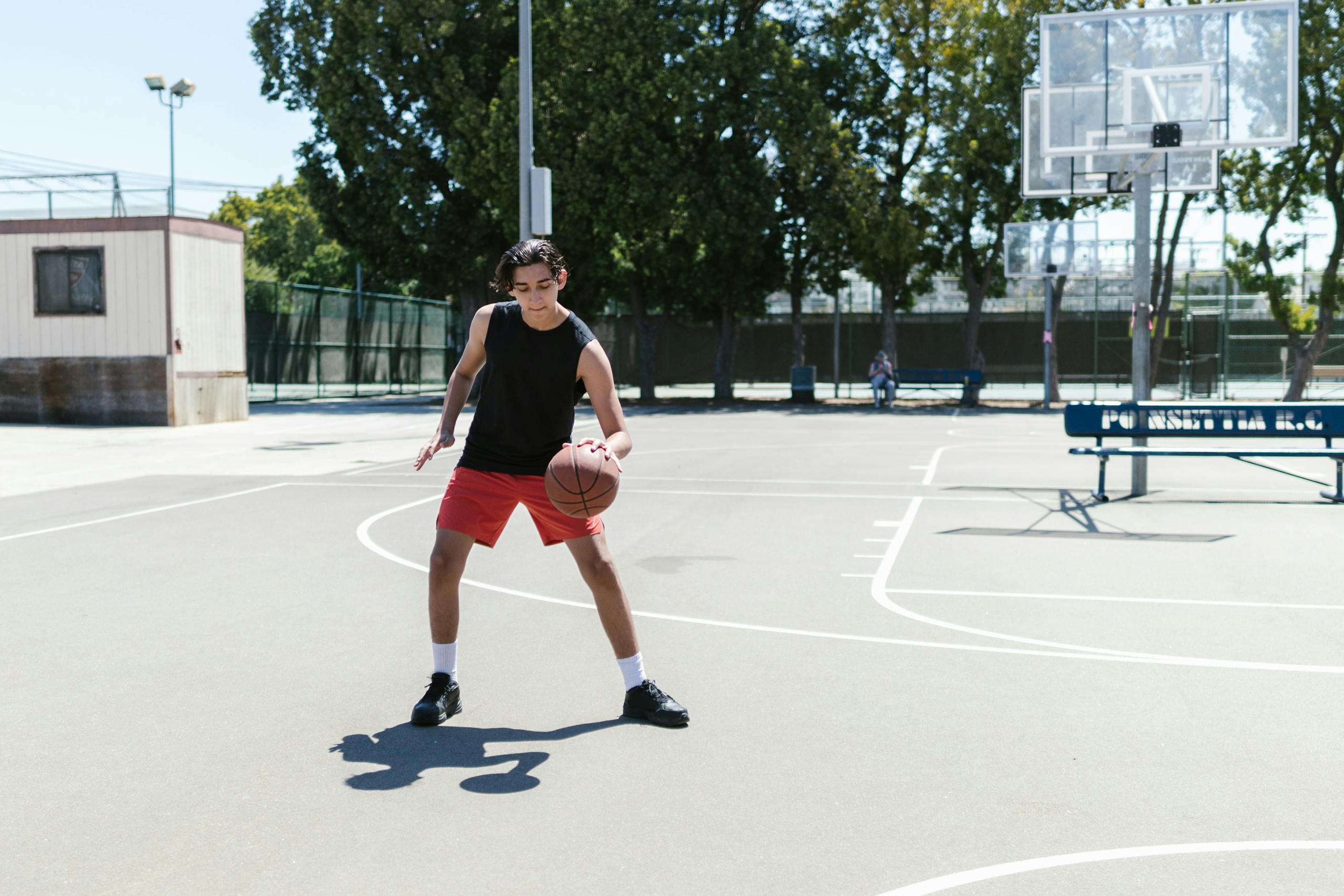 Teenage boy practicing basketball on an outdoor court during a bright summer day.
