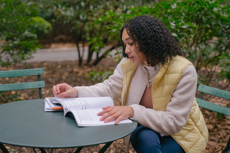 Thoughtful young Hispanic lady in casual clothes sitting in chair at table while reading textbook in park with foliage on ground near green bushes in autumn day