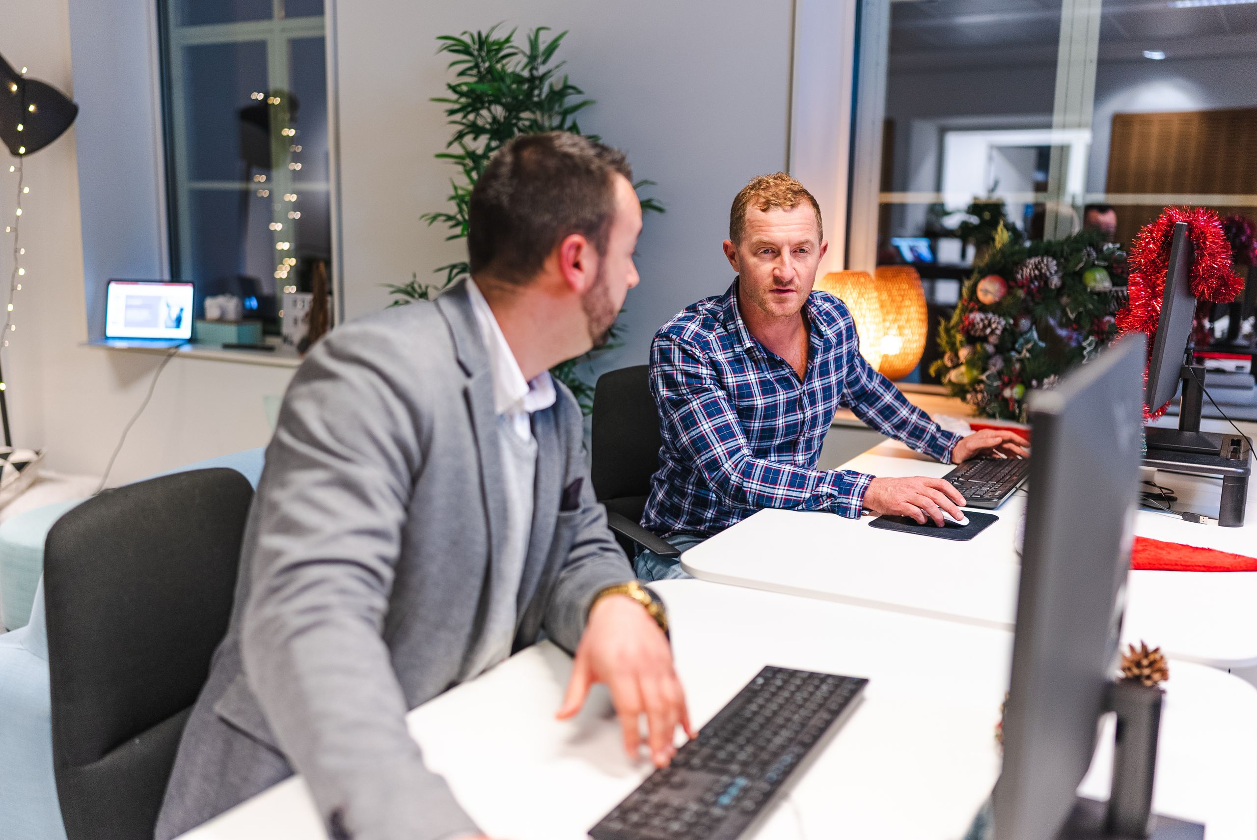 Two men collaborate in a modern office with computers and holiday decor.