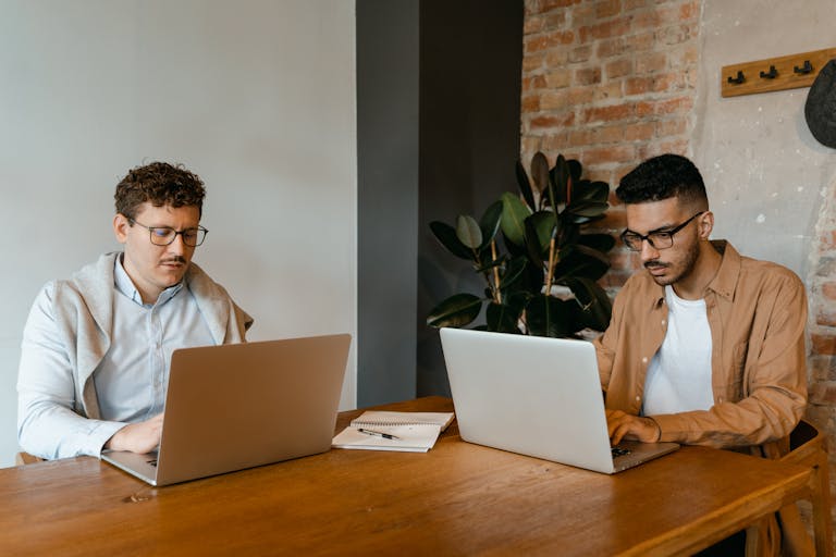 Two men collaborating on laptops indoors at a wooden table, surrounded by a modern brick interior.