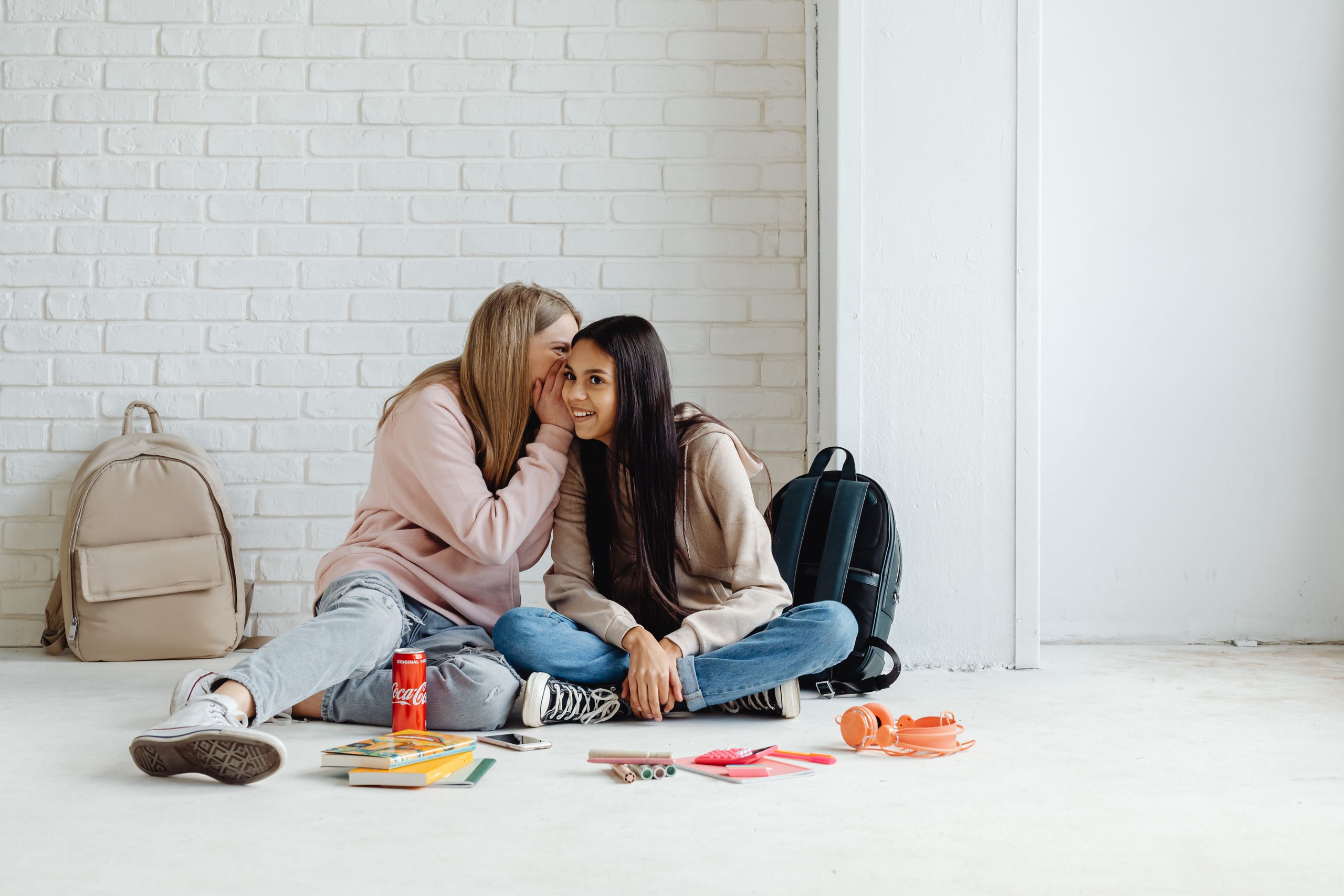 Two teenage girls sitting on the floor, whispering and smiling together.