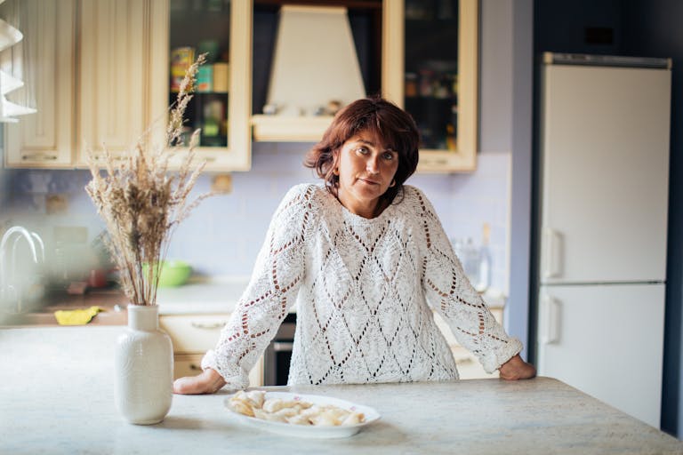 Woman in a kitchen with pelmeni on counter, wearing knitted sweater.