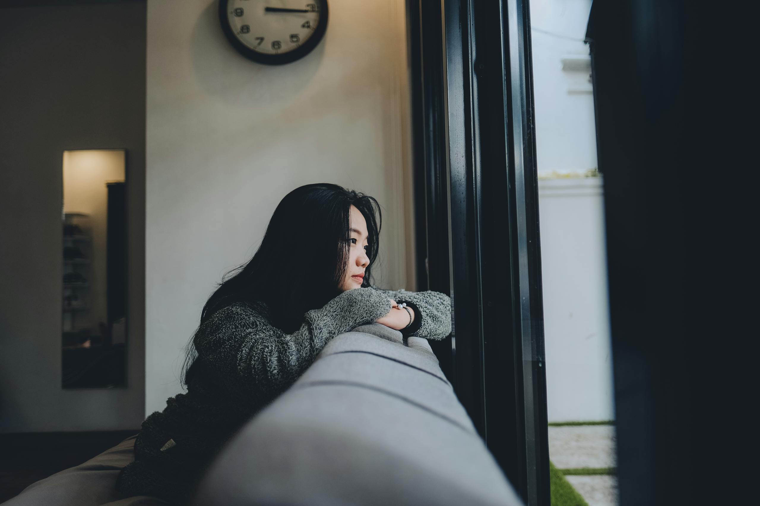Woman leaning on a sofa, looking out of a window in a domestic setting.