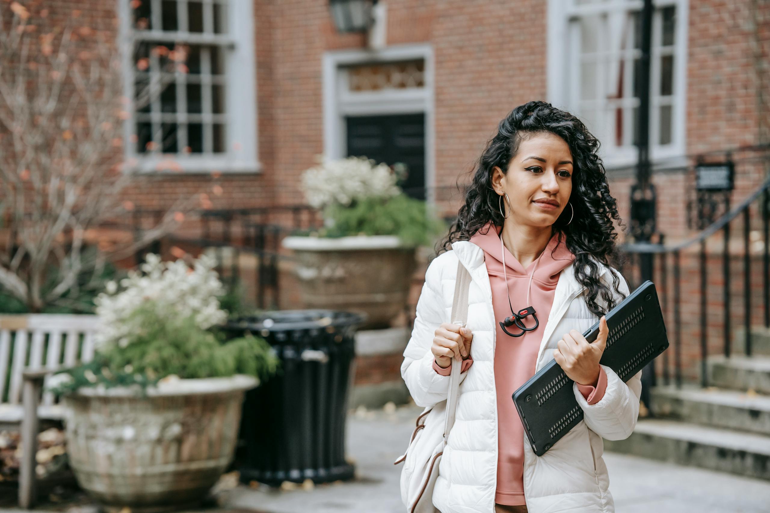 Young ethnic female with long curly hair and netbook in casual trendy outfit on blurred background of brick building