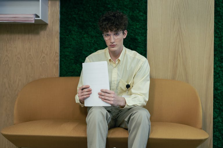 Young man sitting on a sofa in an office holding papers, wearing glasses and a yellow shirt.
