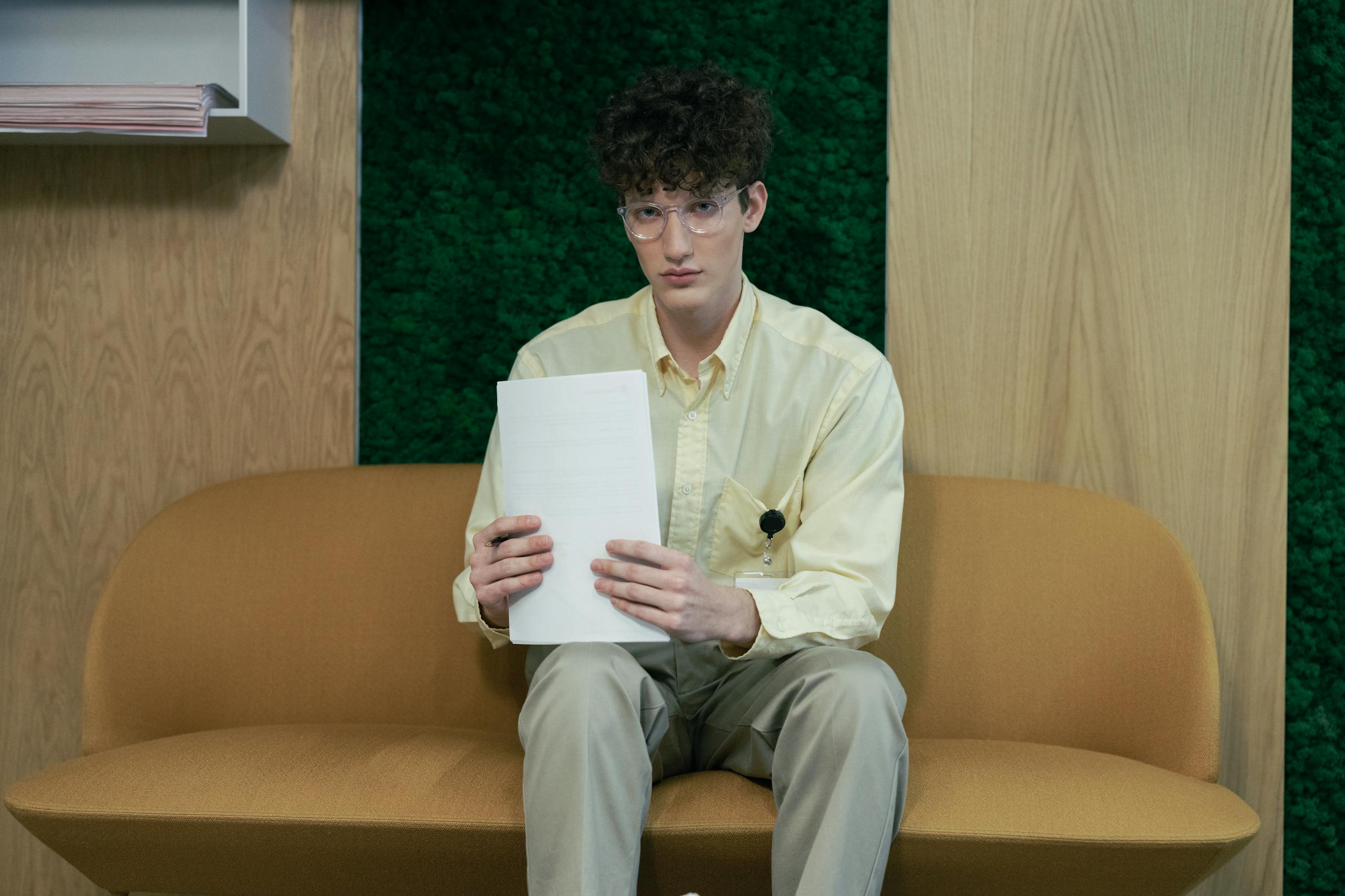 Young man sitting on a sofa in an office holding papers, wearing glasses and a yellow shirt.