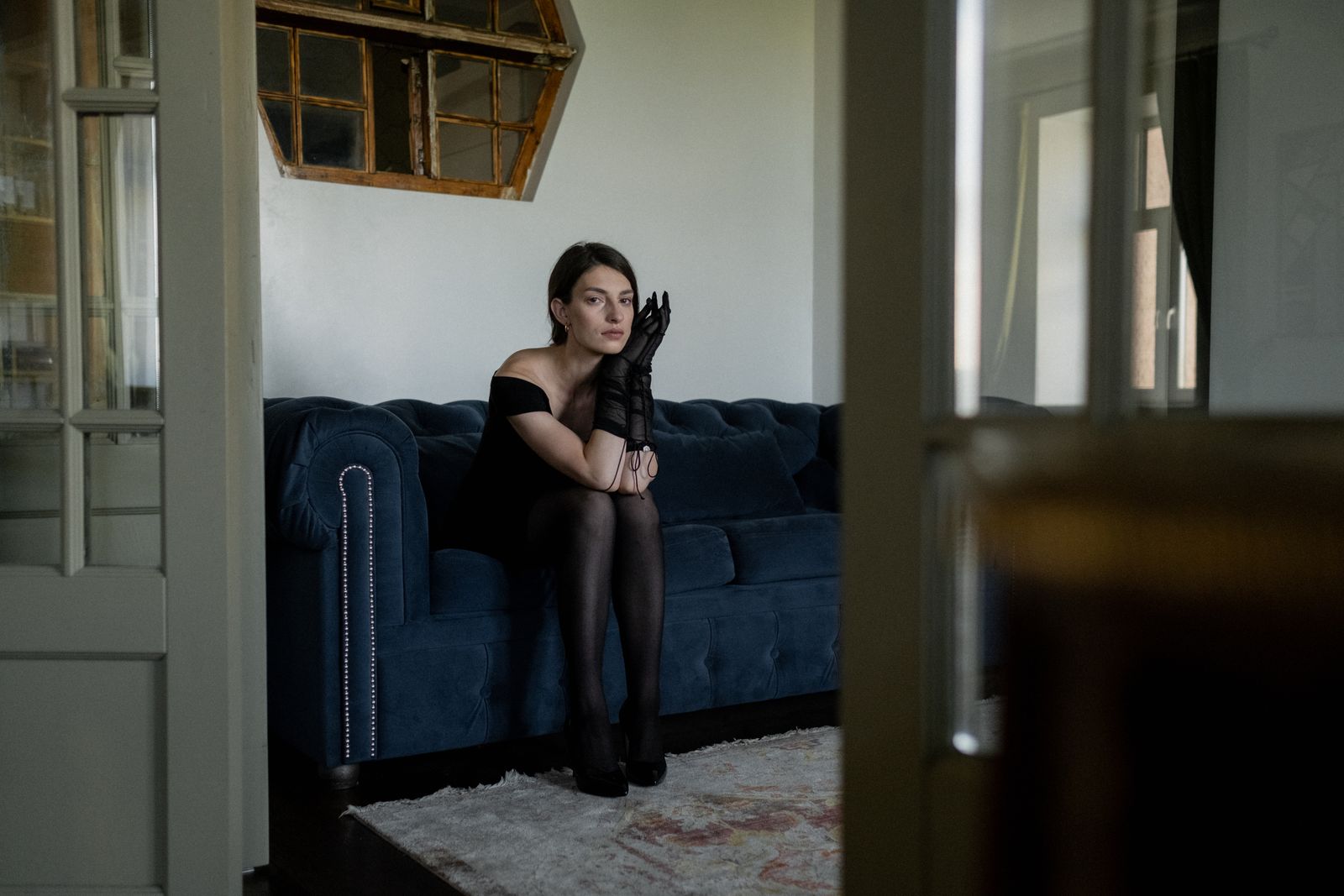 A woman in a black dress sits on a sofa, expressing introspection and sorrow.