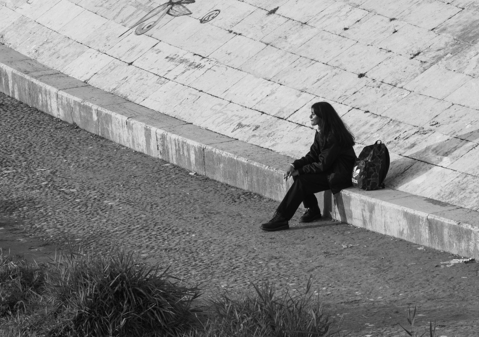 Black and white image of a woman sitting alone by a graffiti-covered wall in an urban setting.