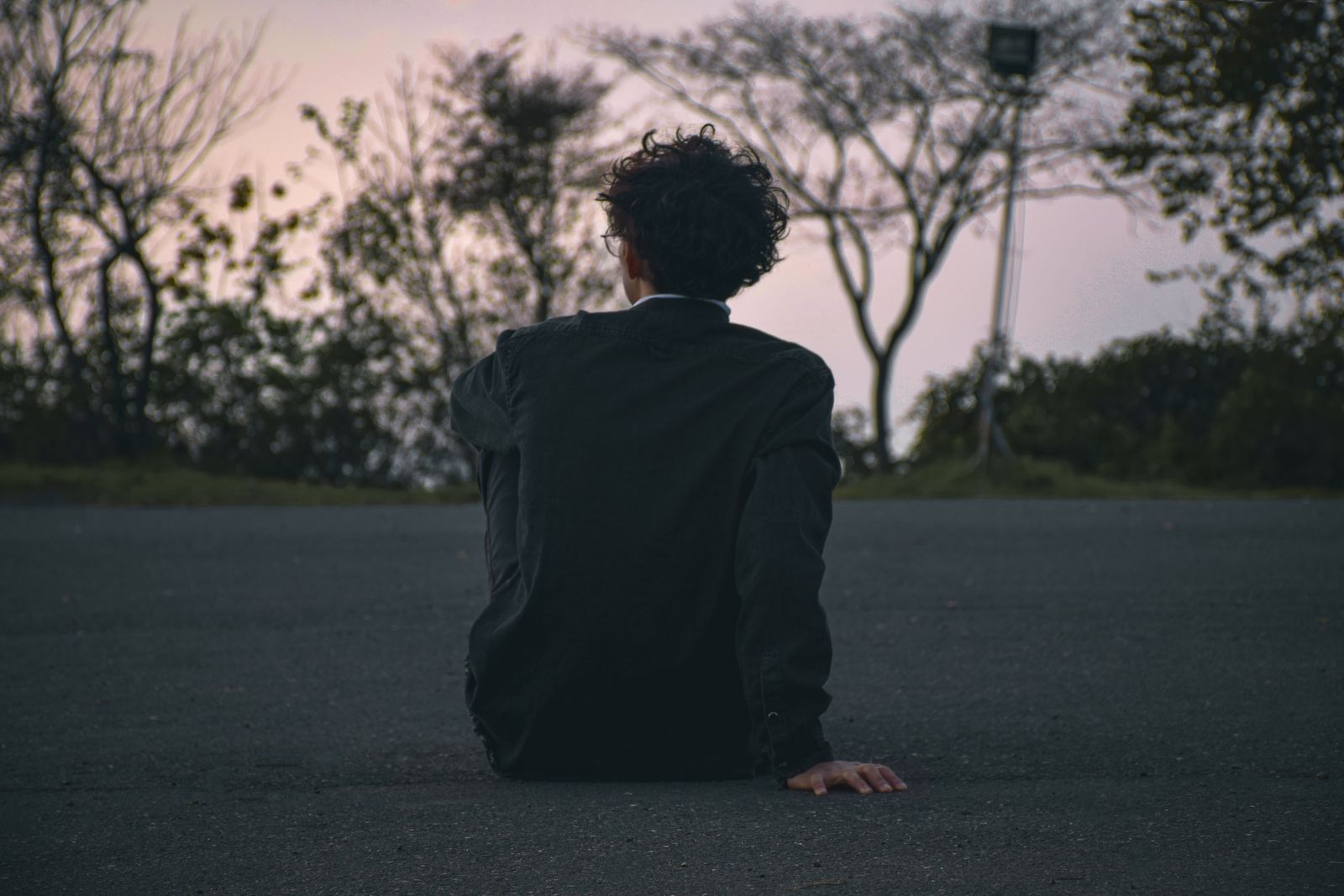 A man with curly hair sits on a road in solitude at dusk, offering a contemplative scene.