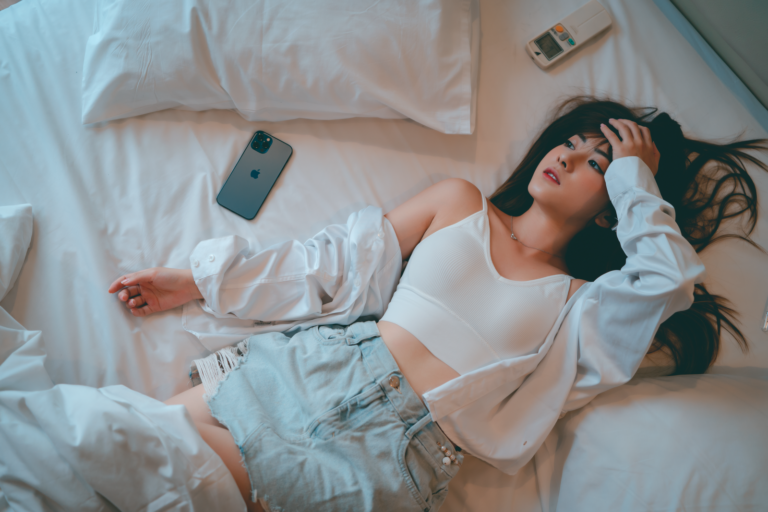 Young woman in casual attire relaxing on bed with a smartphone nearby.