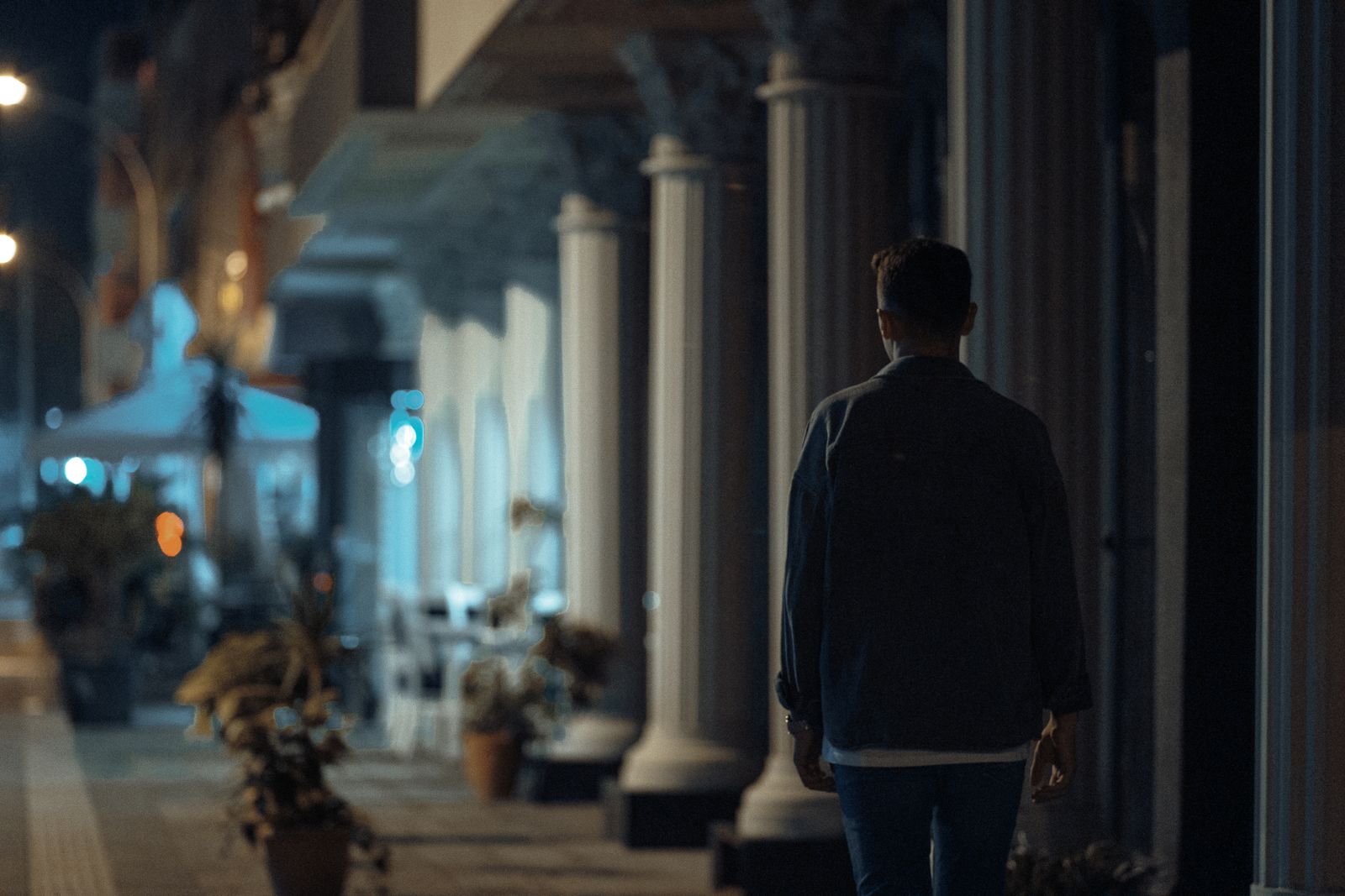 A person walks at night along a city sidewalk with pillars, creating a moody atmosphere.