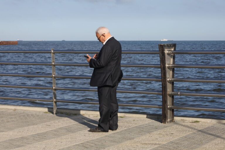 Elderly man in a suit texting on his phone by the ocean. Professional and serene seascape view.