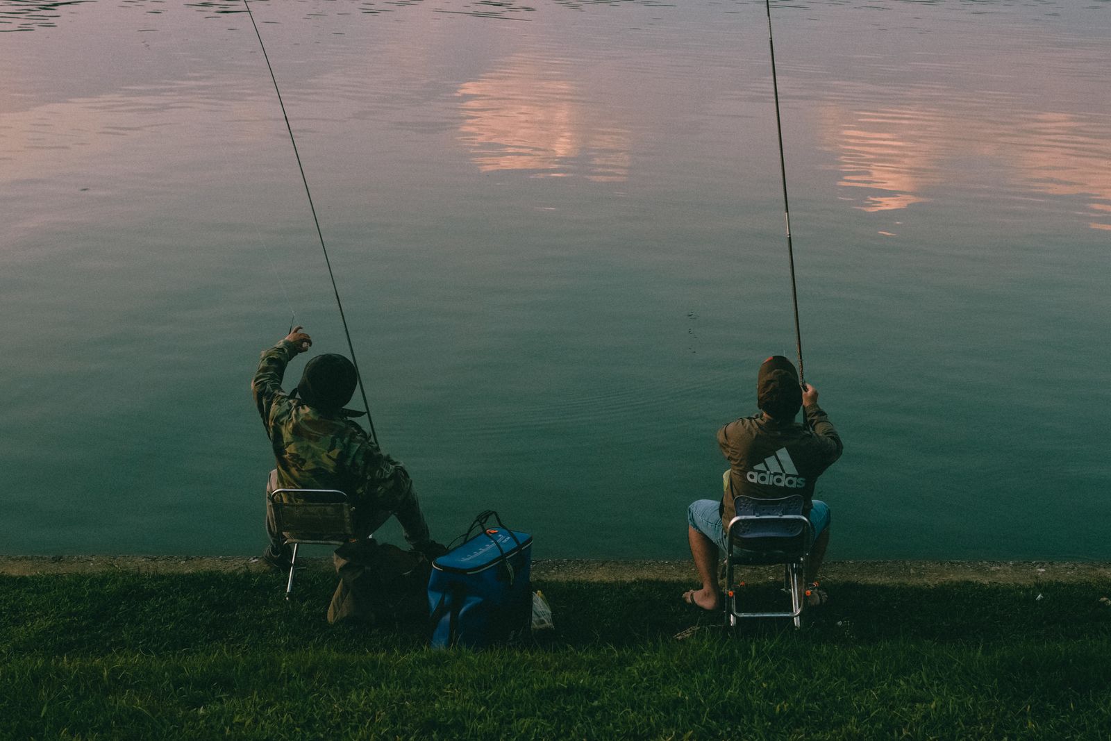 Two people enjoy leisure time fishing by a calm lake during evening hours.