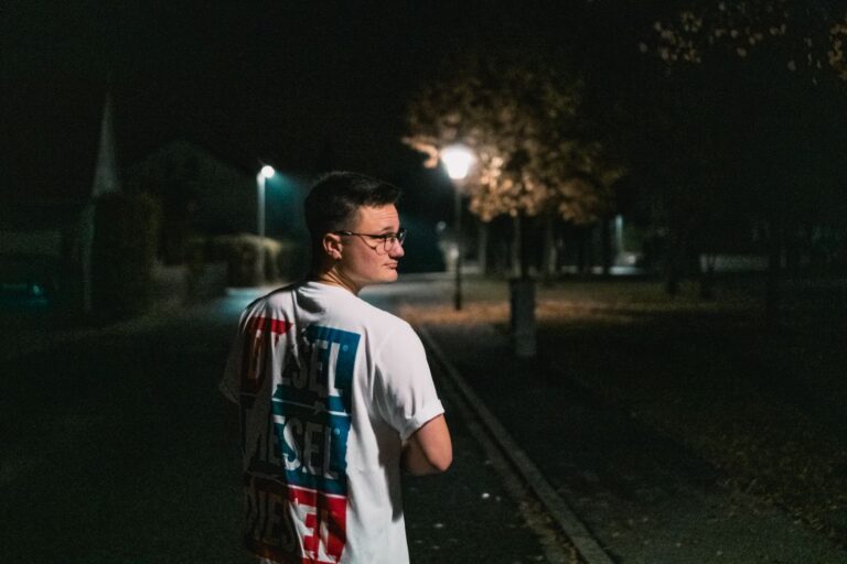 A young man walks on a dimly lit street at night, creating a mysterious urban atmosphere.