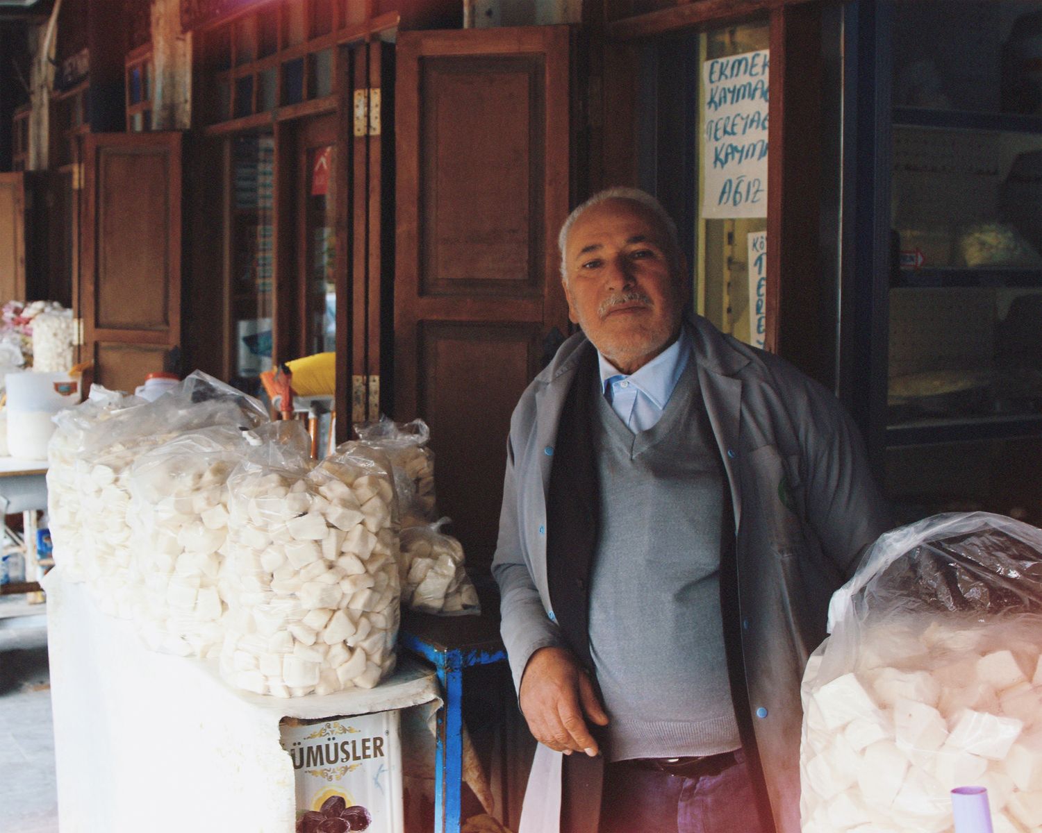 Elderly man standing in a market shop with bags of goods in a traditional setting.
