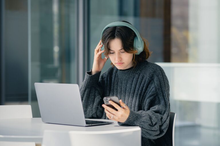Focused young woman with headphones using laptop and smartphone in a modern cafe.