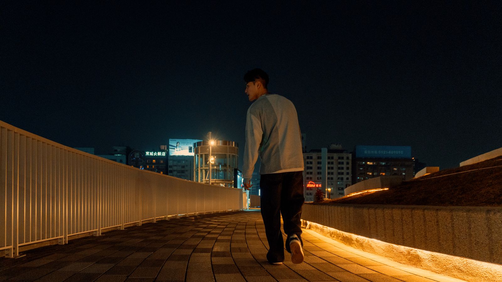 A solitary man walks on a lit city bridge at night, surrounded by a modern urban skyline.