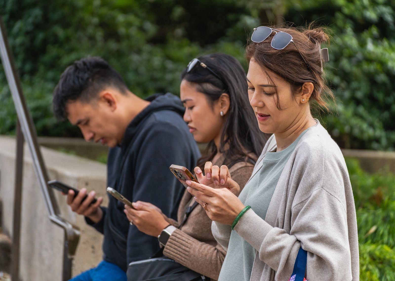 Three young adults focused on their smartphones while sitting outdoors during the day.