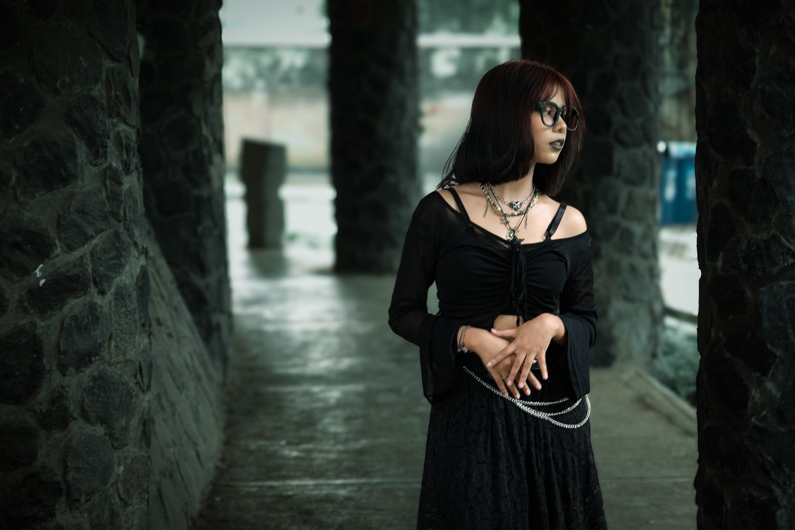 A young adult with Gothic style attire stands thoughtfully in a stone corridor, creating a mysterious atmosphere.