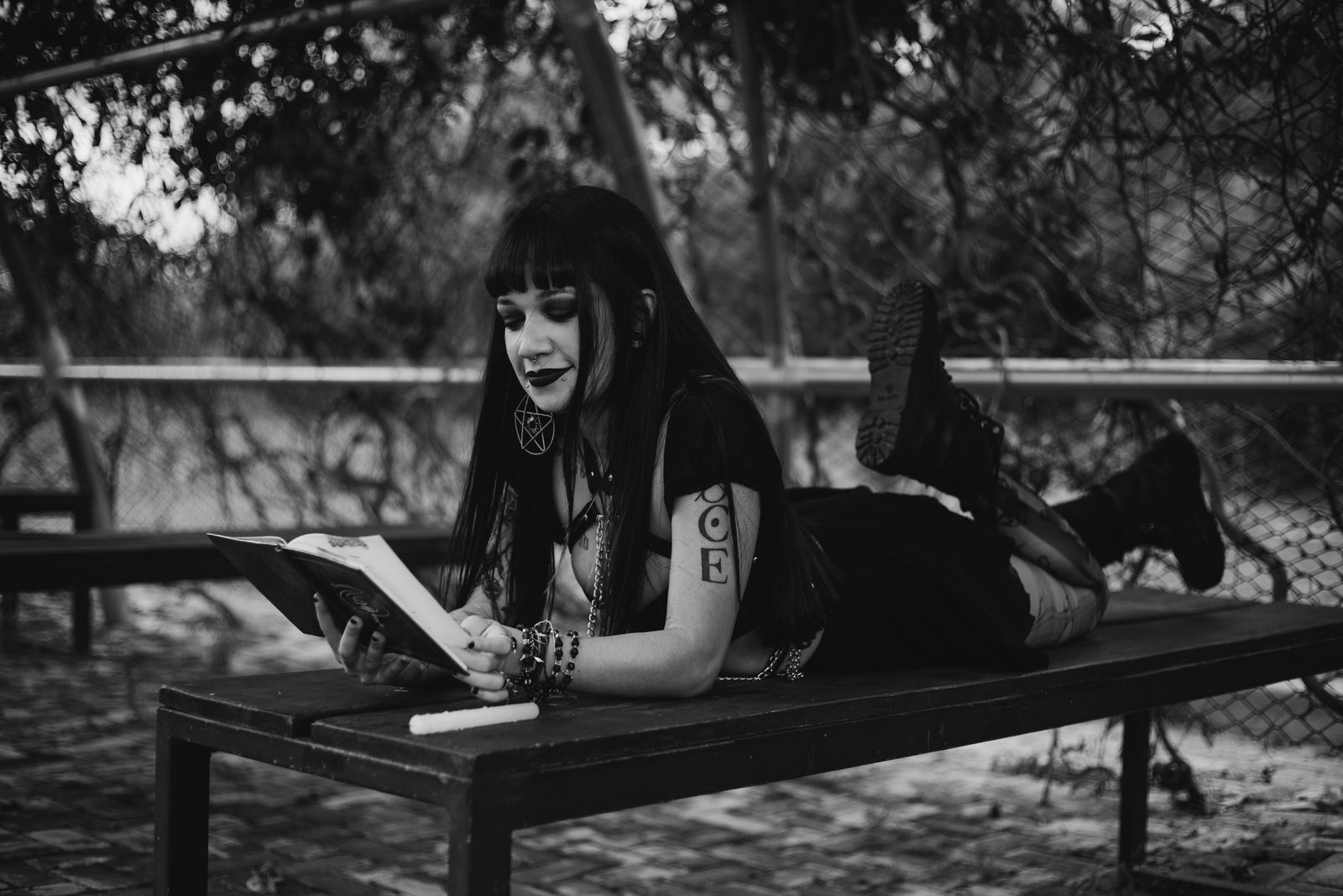 Gothic-themed teenager reading a book outdoors on a bench in a black and white photo.