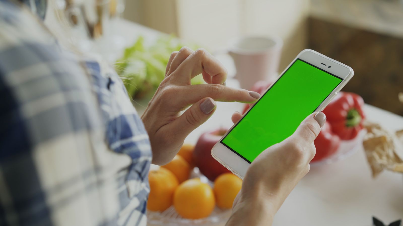 A person using a smartphone with a green screen surrounded by fresh fruits and vegetables in the kitchen.