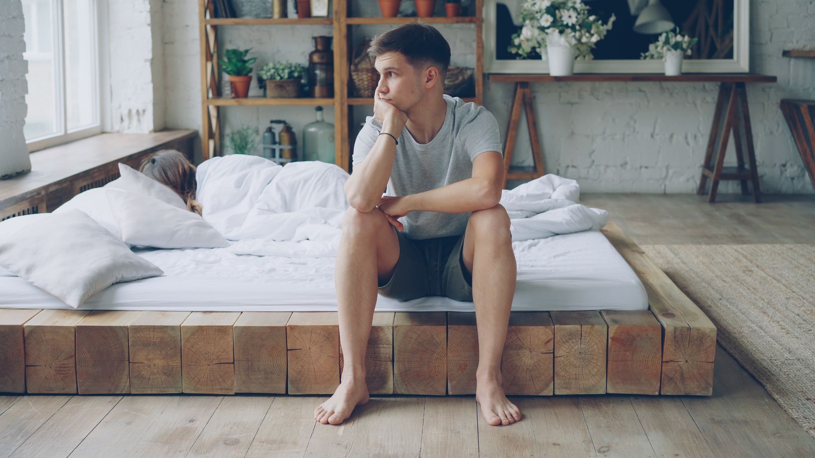 A young man sits thoughtfully on a wooden bed in a cozy, plant-filled bedroom.