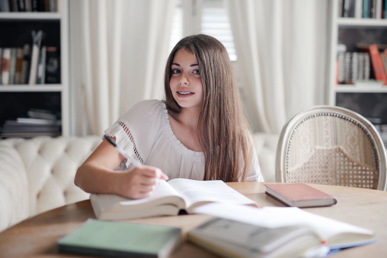 Smiling woman with long hair studying indoors at a table surrounded by books.