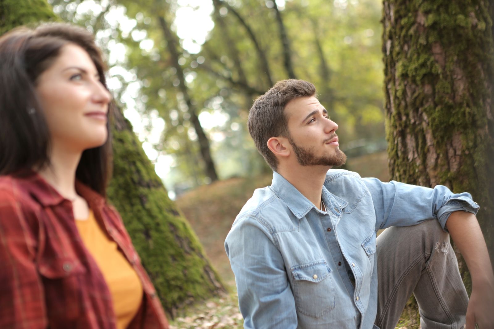Pensive man in denim and blurred woman in red shirt sitting on ground in forest and looking away