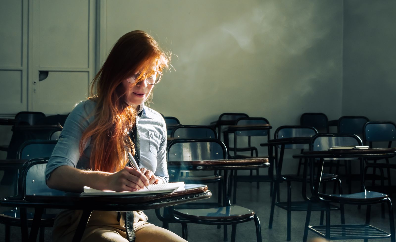 A young woman with red hair studies alone in an empty classroom, sunlight streaming in.