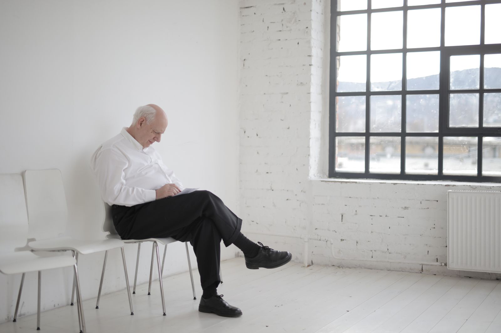 Elderly man in white shirt sitting pensively in a bright, minimalist room with large window.