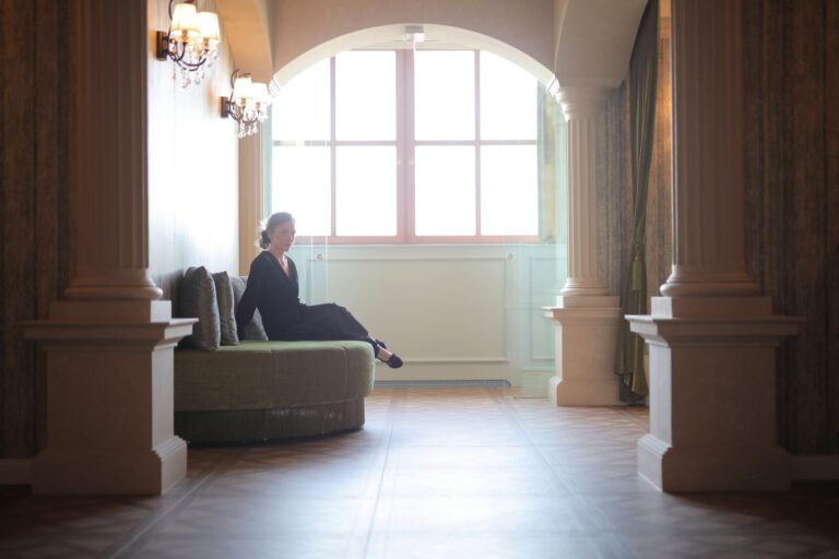 A woman in a black dress sits elegantly in a classic lobby framed by architectural columns.