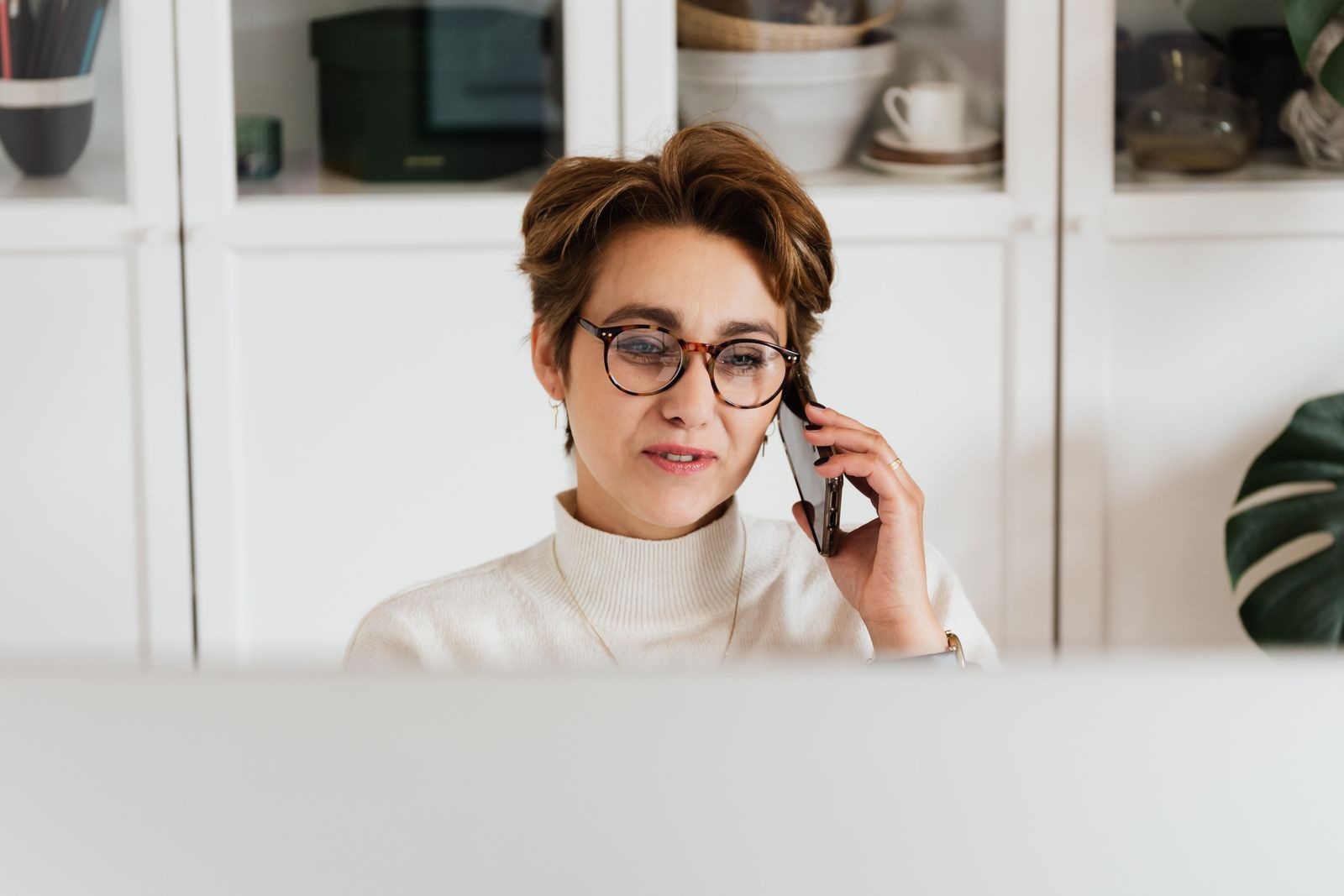 Elegant lady in glasses with short hair discussing current work issues on smartphone while sitting in front of computer in office