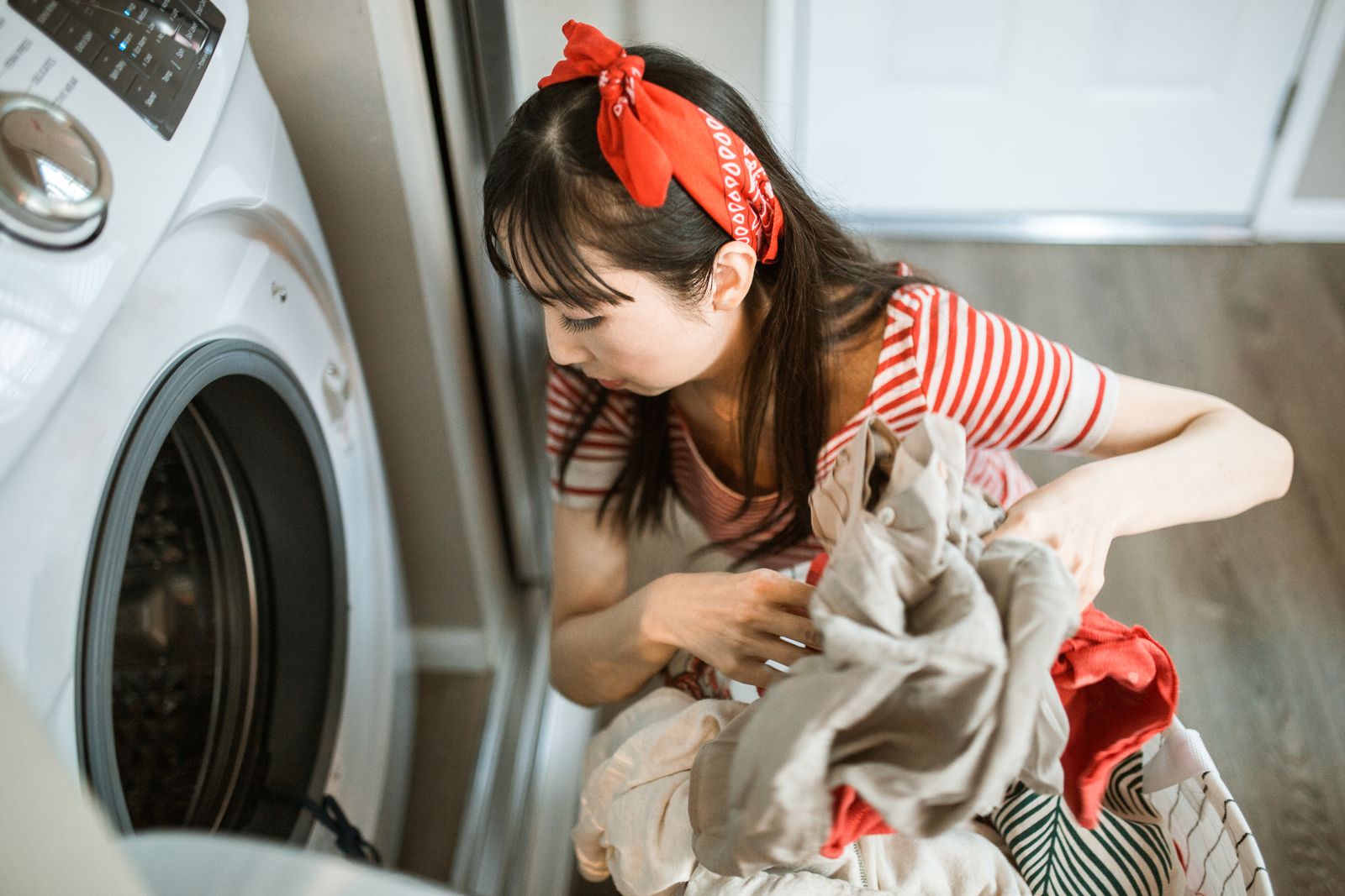 An Asian woman in striped shirt and bandana loading clothes into a washing machine.