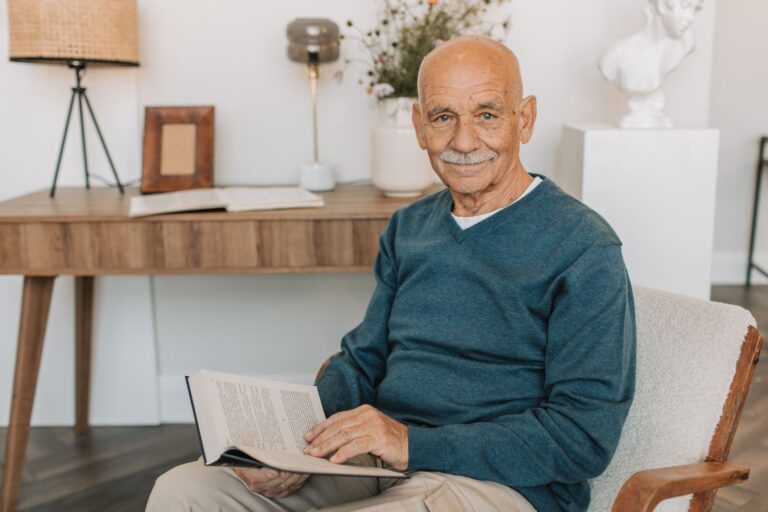 Portrait of a smiling senior man sitting indoors while reading a book for leisure.