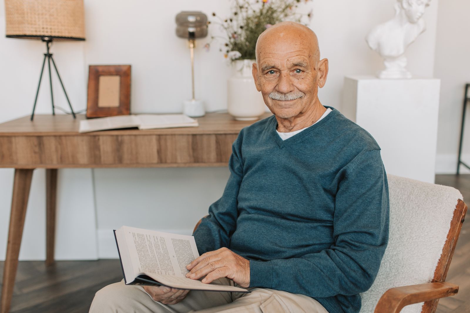 Portrait of a smiling senior man sitting indoors while reading a book for leisure.