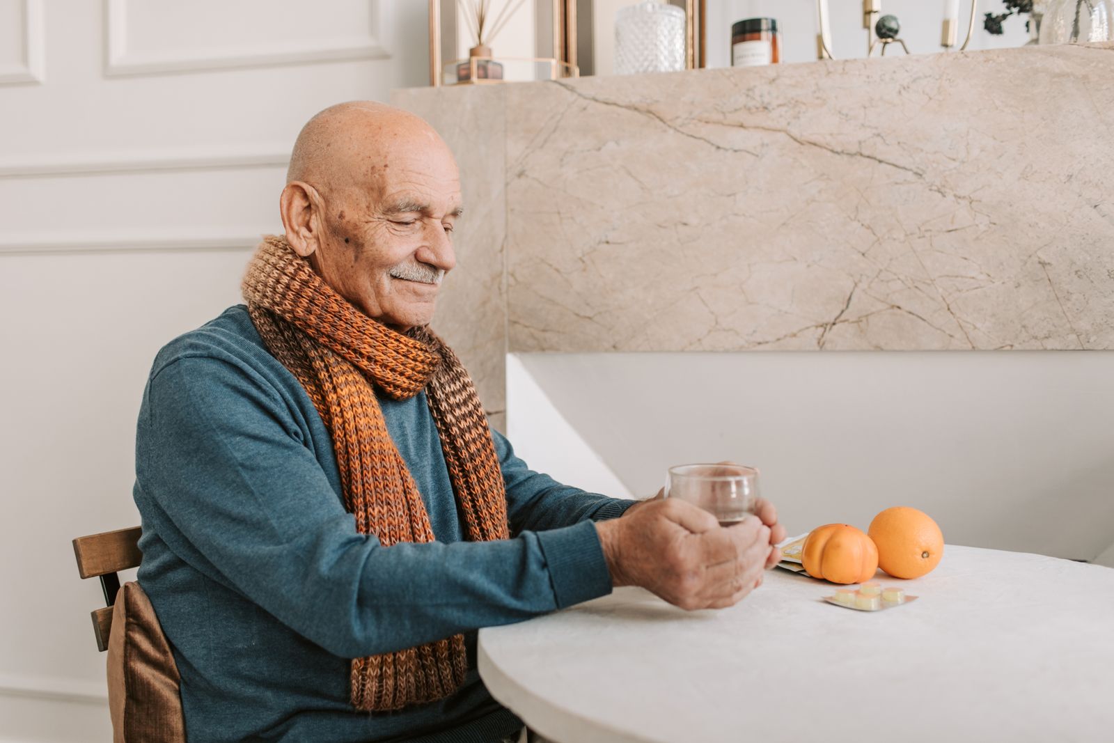 A senior man sitting at a table enjoying a drink, wearing a colorful scarf, with fruits nearby.