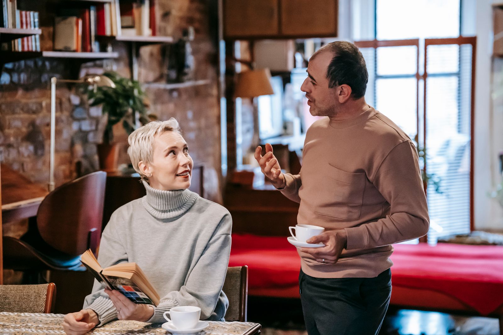 Positive diverse middle aged spouses in casual clothes drinking coffee and discussing interesting book while spending weekend together at home