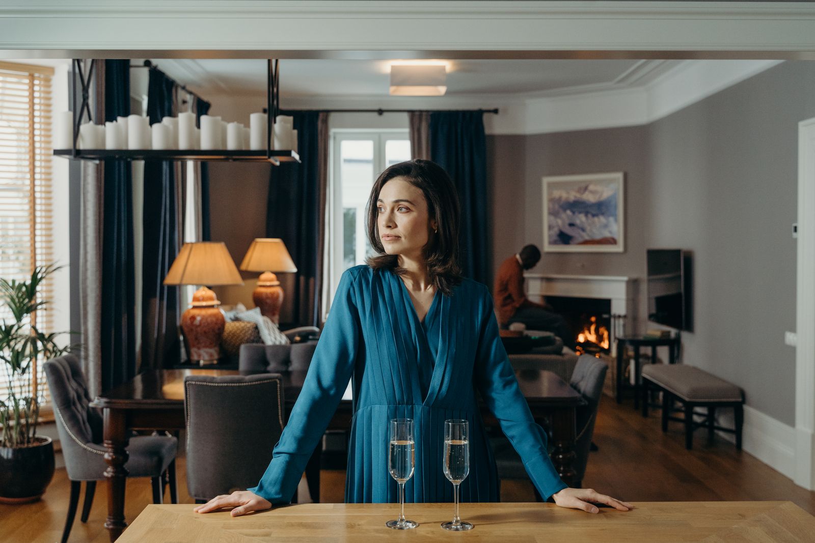 A woman in a blue dress stands by a table with champagne glasses in an elegant living room setting.