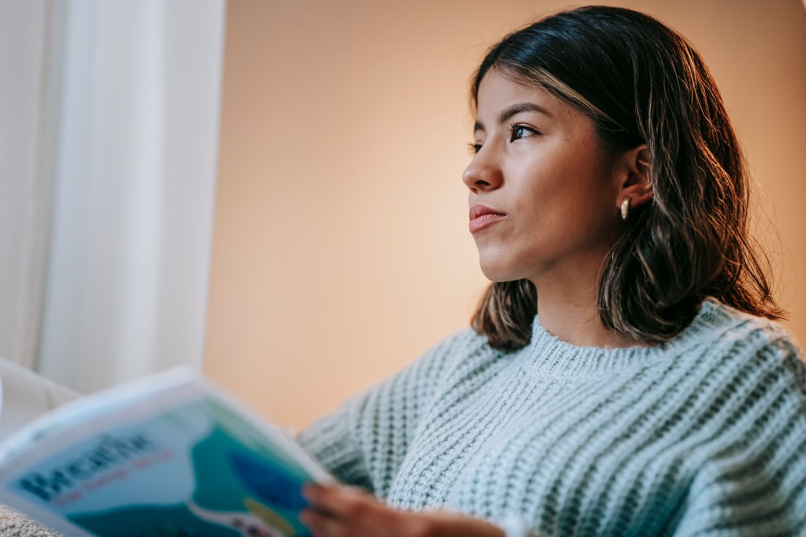 Low angle of pensive Asian female in knitted sweater reading magazine and thoughtfully looking away