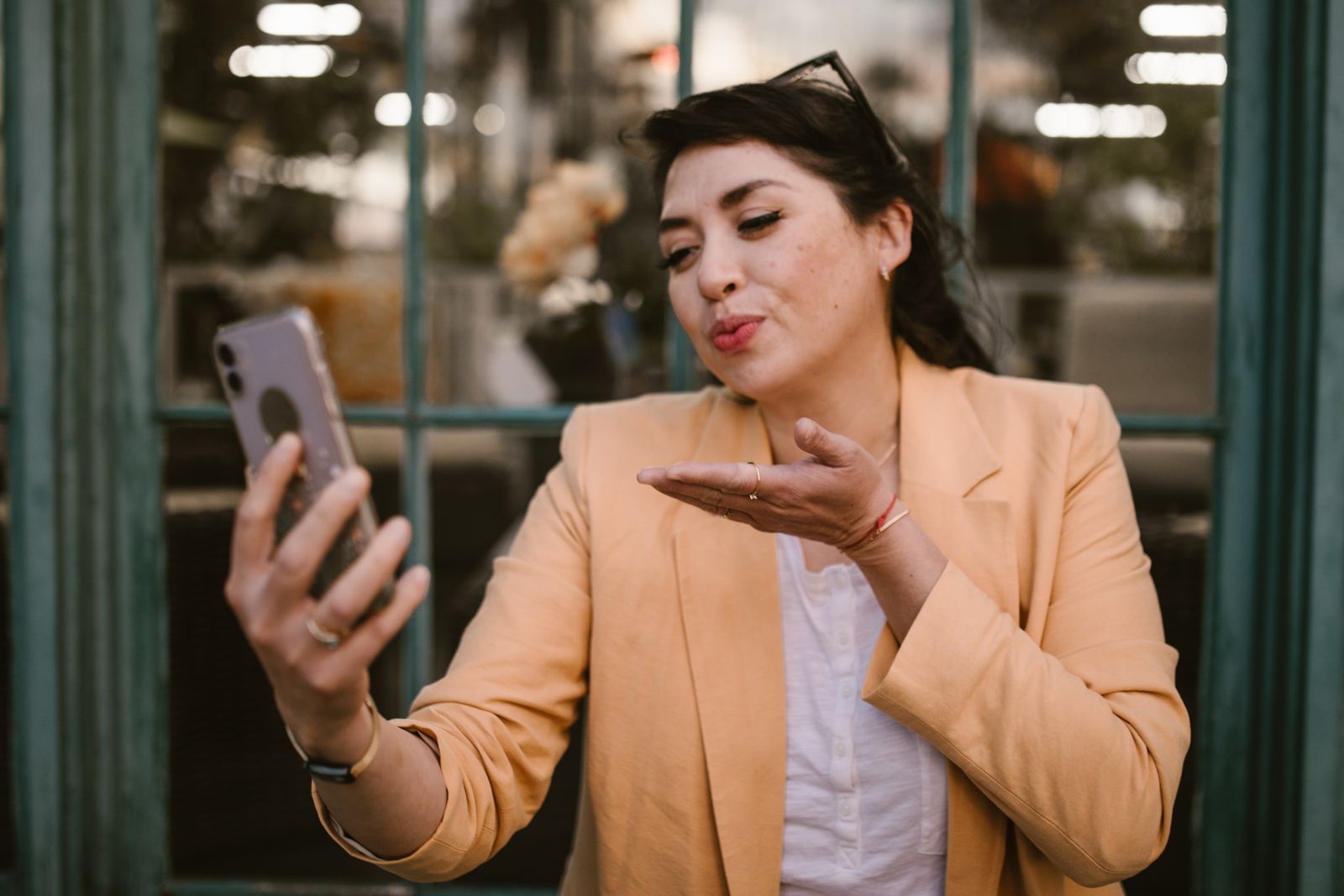 A woman in a beige blazer blows a kiss while talking on a video call outdoors.