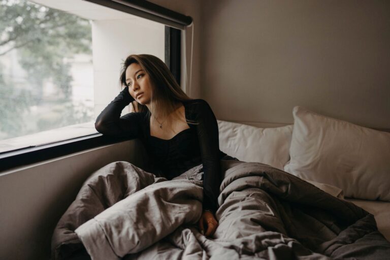 A young woman in a cozy room, pondering by the window, wrapped in blankets.