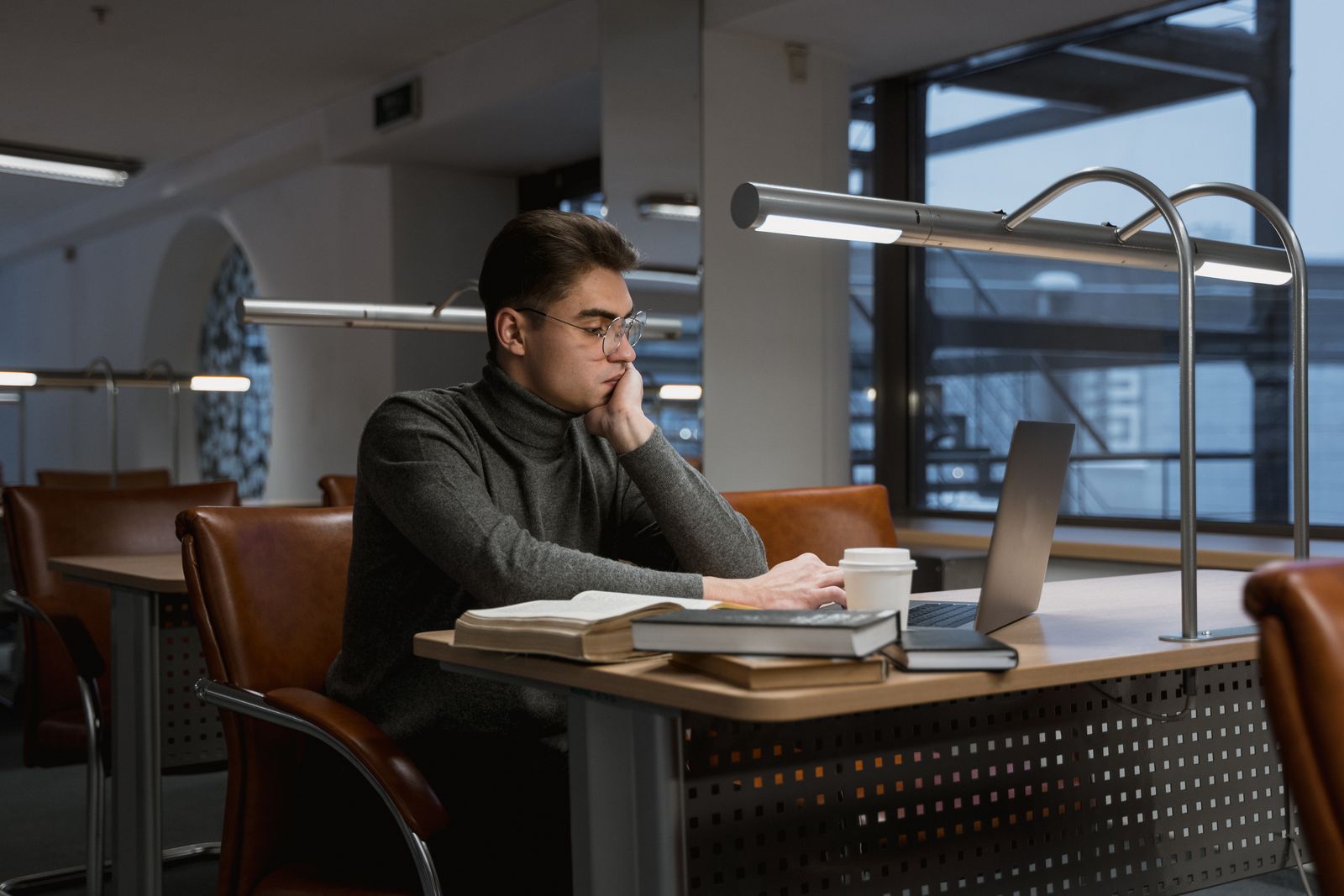 A young man deeply focused while working on a laptop in a quiet, modern library setting.