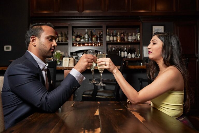Side view of ethnic couple clinking wineglasses with alcohol drink while sitting at table in modern restaurant near bar during romantic date