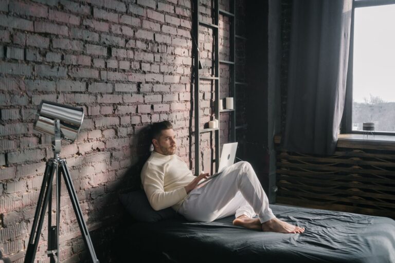 Young man enjoying remote work on laptop in a modern industrial bedroom setting.