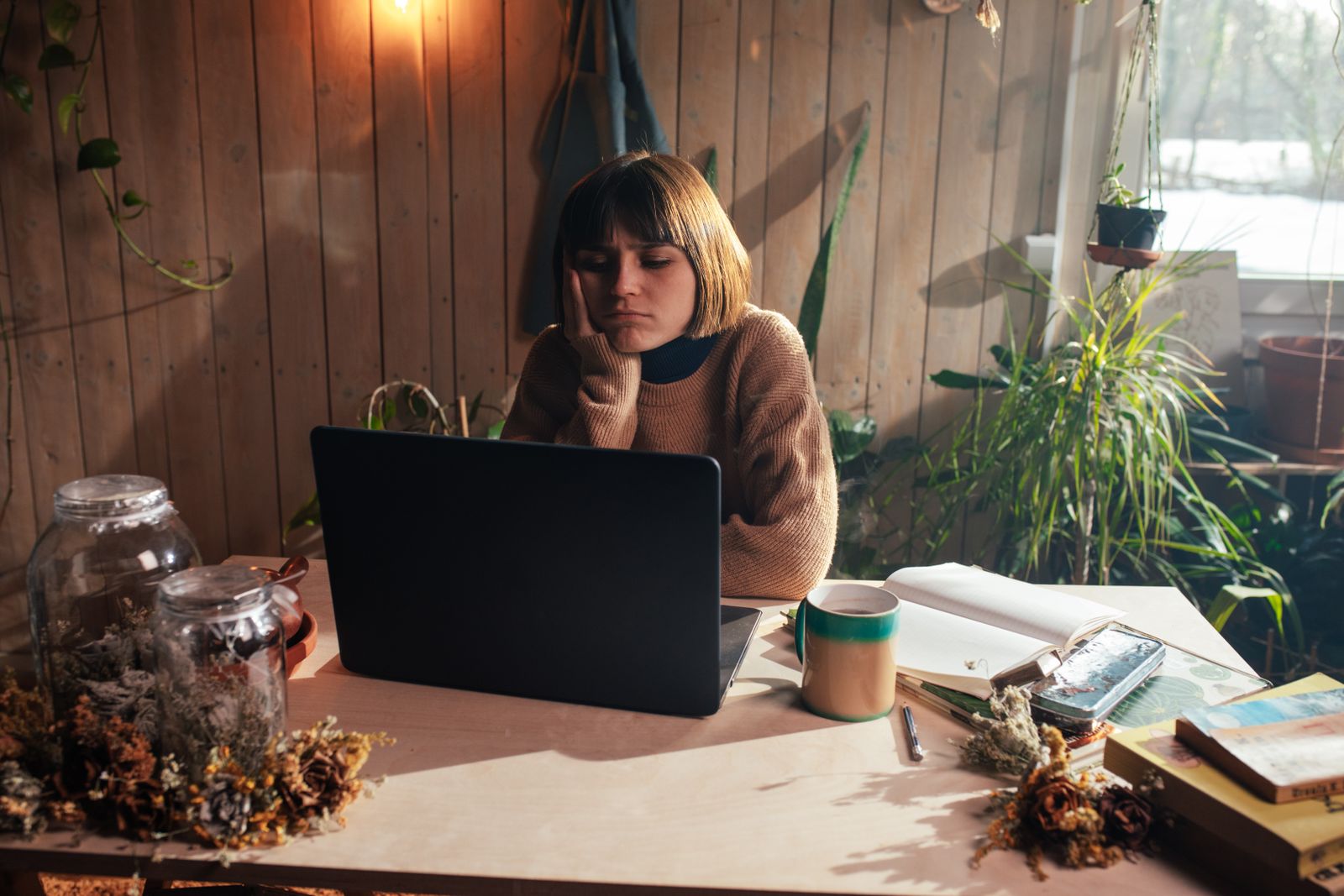 A woman in a cozy room surrounded by plants, appearing bored while using her laptop.