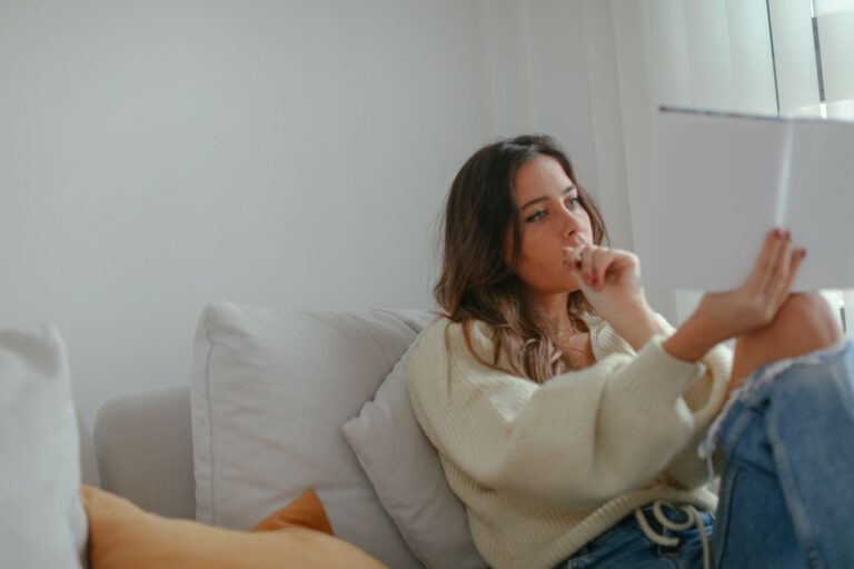 A woman sitting on a couch indoors, thoughtfully reading a book in a cozy setting.